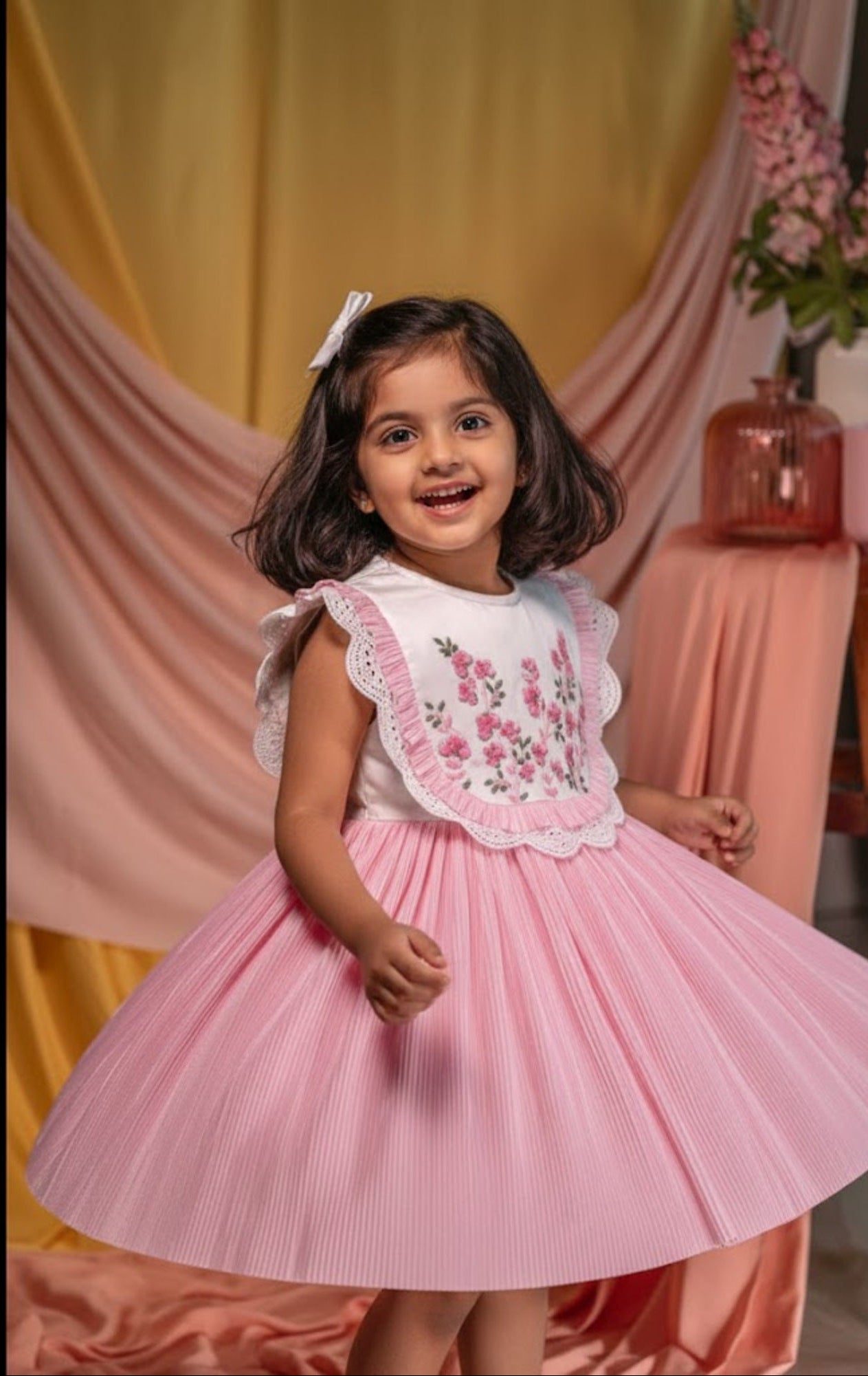 Young girl in a pink dress with floral embroidery standing in front of a yellow and pink draped backdrop.