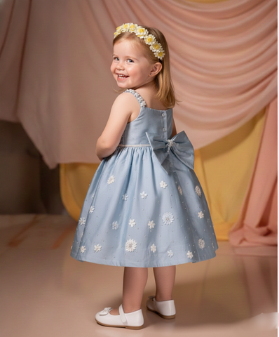 Young girl in a blue floral dress with a bow, standing against a soft pink curtain.